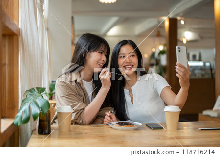 Two beautiful Asian women are enjoying taking pictures together while hanging out at a coffee shop. 118716214