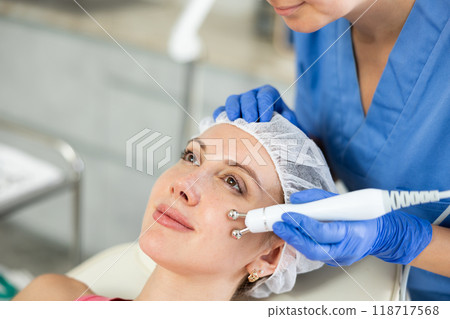 Adult woman cosmetologist doing microcurrent procedure to young woman Adult woman cosmetologist doing microcurrent procedure to young woman 118717568