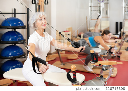 Sports club senior woman client performs exercise on reformer with cable and rope for arms 118717808