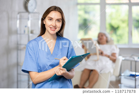 Woman surgeon in uniform is smiling and writing on clipboard in clinic with patient who lying behind Woman surgeon in uniform is smiling and writing on clipboard in clinic with patient who lying behind 118717878