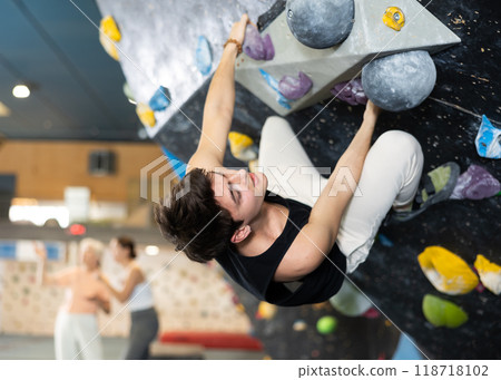 Sporty strong young man exercising in boulder climbing hall without rope, reaching new results, enjoying new challenges in amusement park 118718102