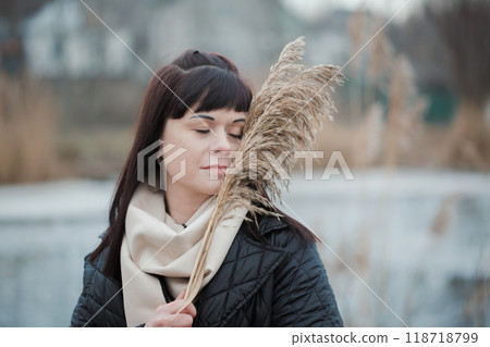 Portrait of young woman wearing coat and scarf with dry miscanthus flower outdoors Portrait of young woman wearing coat and scarf with dry miscanthus flower outdoors 118718799