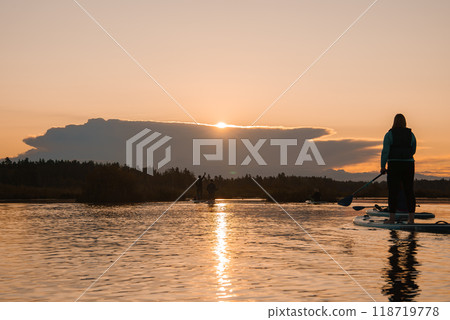 A tranquil scene of people paddleboarding on a calm lake during sunrise. The sun's warm glow and the silhouettes reflected on the water create a picturesque moment. 118719778