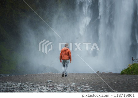 A person in a bright orange jacket and light blue jeans approaches the powerful Skogafoss waterfall in Iceland, surrounded by mist, rocky terrain, and lush green moss. 118719801