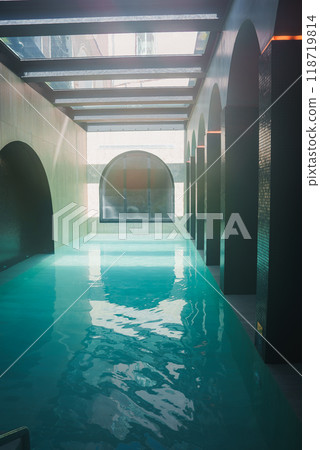 Elegant indoor swimming pool in a high-end luxury hotel, featuring arched openings, skylights, and serene turquoise water illuminated by natural light. 118719814
