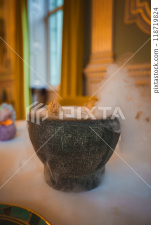 A dark stone bowl emitting misty vapor on a white tablecloth, set in a high-end dining venue with gold accents and tall windows draped with yellow curtains. A dark stone bowl emitting misty vapor on a white tablecloth, set in a high-end dining venue with gold accents and tall windows draped with yellow curtains. 118719824