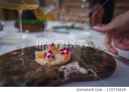 A beautifully presented seafood dish with edible flowers on a dark marble-patterned plate. The elegant table setting and manicured hand suggest a high-end dining experience. A beautifully presented seafood dish with edible flowers on a dark marble-patterned plate. The elegant table setting and manicured hand suggest a high-end dining experience. 118719826