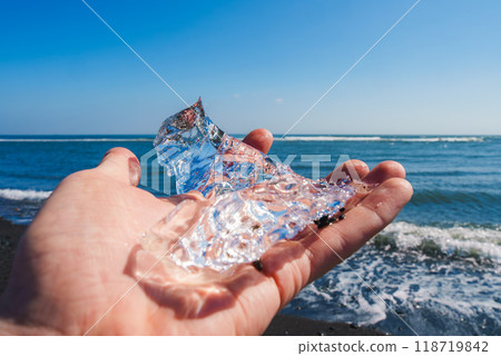 A clear piece of ice is held against a black sand beach and blue ocean on a sunny day in Iceland, showcasing the striking contrast of natural elements. A clear piece of ice is held against a black sand beach and blue ocean on a sunny day in Iceland, showcasing the striking contrast of natural elements. 118719842
