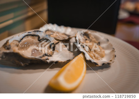A detailed view of fresh oysters with a lemon wedge, set against a blurred, elegant interior. The warm lighting and meticulous presentation highlight a high-end dining experience. 118719850