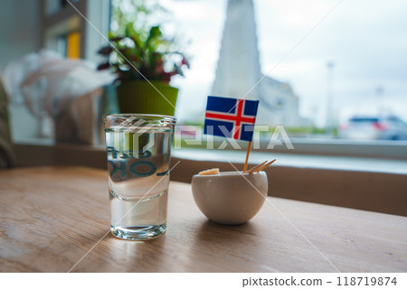 A small table setup inside a cafe featuring a glass of clear liquid and a bowl with an Icelandic flag. Hallgrimskirkja church is visible through the window. A small table setup inside a cafe featuring a glass of clear liquid and a bowl with an Icelandic flag. Hallgrimskirkja church is visible through the window. 118719874