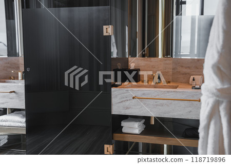 Elegant hotel bathroom featuring a sleek black glass door, marble countertop, gold fixtures, and neatly folded towels on a wooden shelf beneath the sink. Elegant hotel bathroom featuring a sleek black glass door, marble countertop, gold fixtures, and neatly folded towels on a wooden shelf beneath the sink. 118719896