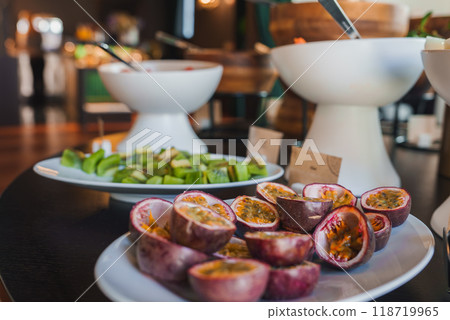 Close-up of a plate with halved passion fruits and sliced kiwi at a luxurious buffet. Elegant white bowls and warm lighting enhance the sophisticated decor. 118719965