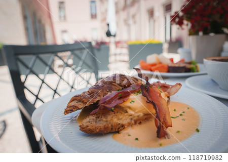 A beautifully plated breakfast featuring crispy bacon, a croissant, and creamy sauce on an elegant white plate. Set in an upscale outdoor dining area with stylish decor. A beautifully plated breakfast featuring crispy bacon, a croissant, and creamy sauce on an elegant white plate. Set in an upscale outdoor dining area with stylish decor. 118719982