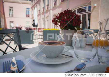 Beautifully set outdoor dining area featuring cappuccino on white tablecloth, elegant tableware, and charming courtyard with pastel pink buildings and red flowers. Beautifully set outdoor dining area featuring cappuccino on white tablecloth, elegant tableware, and charming courtyard with pastel pink buildings and red flowers. 118719994