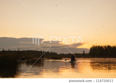 A serene scene of people on SUP boards watching the sunrise over a calm lake, surrounded by dense forest. The sky is painted with soft hues, reflecting on the water. 118720011