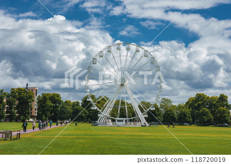 A spacious park in Cambridge, UK, featuring a prominent Ferris wheel, lush trees, and a partly cloudy sky. People walk along a pathway with benches. A spacious park in Cambridge, UK, featuring a prominent Ferris wheel, lush trees, and a partly cloudy sky. People walk along a pathway with benches. 118720019