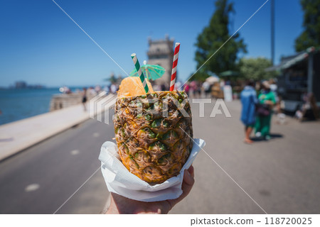A vibrant scene featuring a hand holding a pineapple drink with two straws and a small umbrella, set against the iconic Belem Tower in Lisbon, Portugal. A vibrant scene featuring a hand holding a pineapple drink with two straws and a small umbrella, set against the iconic Belem Tower in Lisbon, Portugal. 118720025