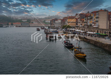 Traditional Rabelo boats moored along the waterfront with colorful historic buildings in Porto's Ribeira district. A warm sunset glow enhances the vibrant scene. Traditional Rabelo boats moored along the waterfront with colorful historic buildings in Porto's Ribeira district. A warm sunset glow enhances the vibrant scene. 118720026