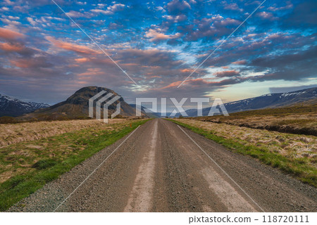 A gravel road stretches into the distance, flanked by grassy fields and leading towards a prominent, rounded mountain under a vibrant, partly cloudy sky in Iceland. A gravel road stretches into the distance, flanked by grassy fields and leading towards a prominent, rounded mountain under a vibrant, partly cloudy sky in Iceland. 118720111