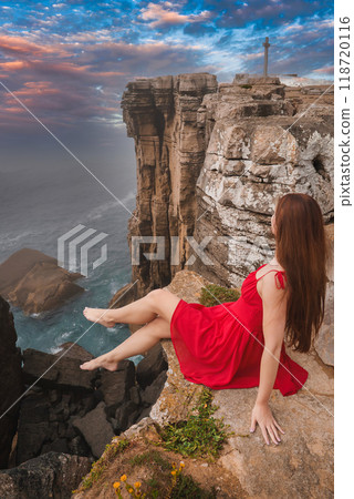 A woman in a red dress sits on a cliff edge, overlooking rugged cliffs and crashing ocean waves at sunset. A tall cross-like monument stands in the background, Portugal. 118720116