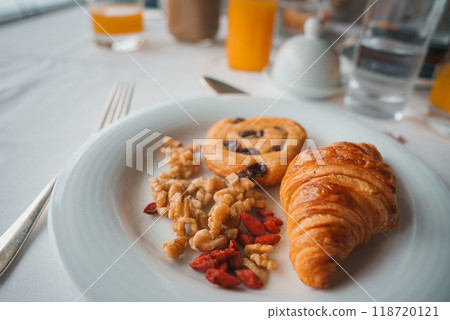 A beautifully arranged breakfast plate featuring a croissant and pastry with raisins, walnuts, and goji berries. Set on a white tablecloth with silver cutlery and glasses of orange juice and water. A beautifully arranged breakfast plate featuring a croissant and pastry with raisins, walnuts, and goji berries. Set on a white tablecloth with silver cutlery and glasses of orange juice and water. 118720121