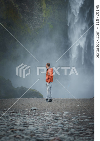 A person in an orange jacket stands on rocky ground, admiring the powerful Skogafoss waterfall in Iceland. The misty atmosphere and moss-covered cliff enhance the scene. 118720149