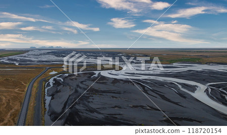 A stunning aerial view of a wide, shallow river winding through black sand in Iceland. A road runs along the left side, with a blue sky and white clouds above. 118720154