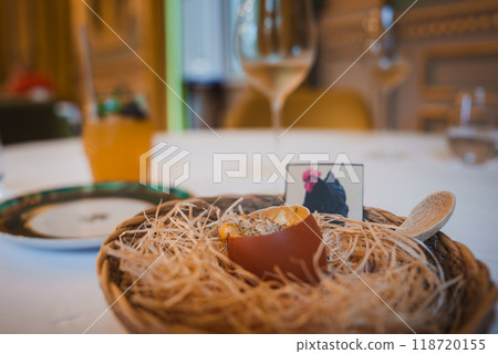 Elegant dining scene featuring a unique dish served in a halved eggshell on straw, with a rooster card, wooden spoon, and fine decor in a luxury hotel. 118720155