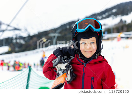 Portrait of cute confident adorable little kid boy enjoy having fun walking with ski at alpine mountain skiing resort on bright winter day. Toddler beginner skier rest of training in ski school Portrait of cute confident adorable little kid boy enjoy having fun walking with ski at alpine mountain skiing resort on bright winter day. Toddler beginner skier rest of training in ski school 118720160
