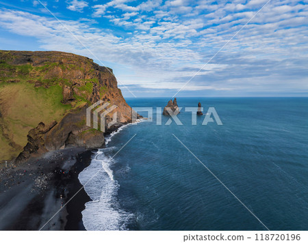 Stunning aerial view of Iceland's rugged coastline featuring a black sand beach, rocky cliff, and distant sea stacks under a bright blue sky with white clouds. 118720196