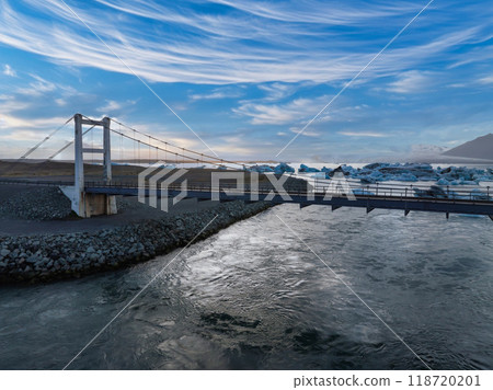 A metal suspension bridge with a wooden walkway spans a wide dark blue river in Iceland. The bridge is surrounded by a rocky shore under a blue sky with clouds. A metal suspension bridge with a wooden walkway spans a wide dark blue river in Iceland. The bridge is surrounded by a rocky shore under a blue sky with clouds. 118720201