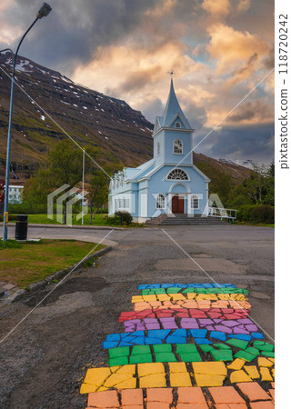 The picturesque blue Seydisfjordur Church in Iceland, set against snow-capped mountains and a dramatic sky, with a vibrant rainbow cobblestone path leading up to it. The picturesque blue Seydisfjordur Church in Iceland, set against snow-capped mountains and a dramatic sky, with a vibrant rainbow cobblestone path leading up to it. 118720242