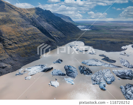 A stunning aerial view of a glacial lagoon in Iceland, featuring milky blue water filled with large ice chunks, surrounded by a barren, rocky landscape and a mountain. 118720252