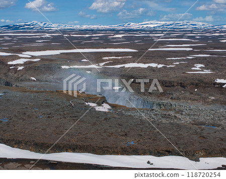 A breathtaking aerial view of Iceland's snow-covered landscape featuring a river with mist, small waterfalls, and distant hills and mountains. 118720254