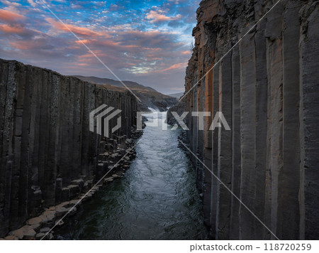 A stunning aerial view of a river winding through a narrow canyon with tall, dark rock formations. The sky above features a beautiful mix of blue and pink clouds. A stunning aerial view of a river winding through a narrow canyon with tall, dark rock formations. The sky above features a beautiful mix of blue and pink clouds. 118720259
