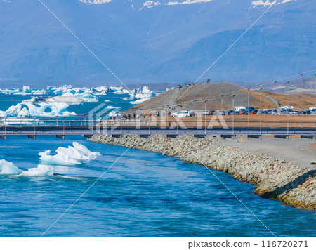 A metal bridge with a wooden walkway spans a bright blue river with floating icebergs and a backdrop of snow-covered mountains in Iceland. A metal bridge with a wooden walkway spans a bright blue river with floating icebergs and a backdrop of snow-covered mountains in Iceland. 118720271