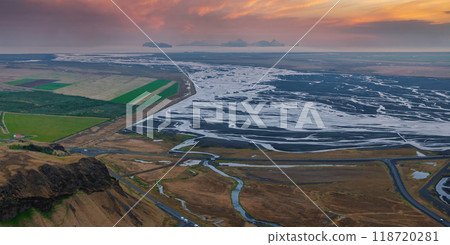 Stunning aerial view of a vast, winding river in Iceland, surrounded by a mix of brown, green, and black terrain. Small buildings are visible in the distance. 118720281