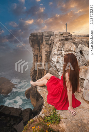 A woman in a red dress sits on a rugged cliff at Cabo da Roca, Portugal, overlooking the dramatic coastal landscape and a prominent cross at sunset. 118720283