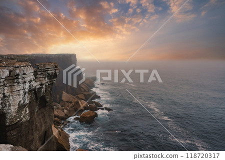 Breathtaking view of the western coast of Portugal featuring dramatic cliffs, vibrant sunset hues, and misty ocean spray. Waves crash against the rugged rocks. 118720317