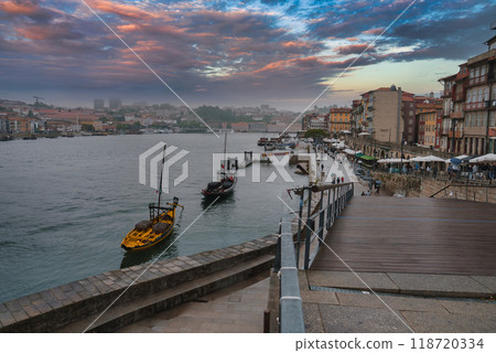 Vibrant view of Ribeira district in Porto with Rabelo boats, colorful buildings, and Dom Luis I Bridge at sunset. People enjoy the lively riverside atmosphere. 118720334