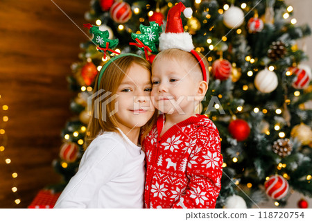 Young boy and girl with playful holiday headbands are standing beside the Christmas tree. 118720754