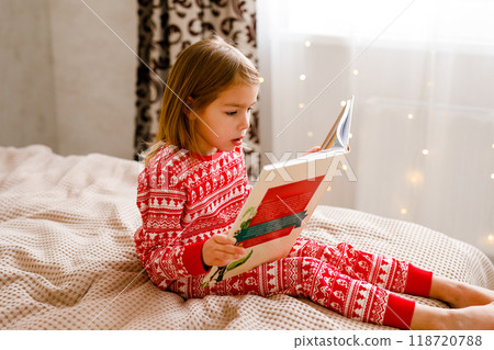 A little girl wearing holiday sleepwear is on the bed, reading a big book 118720788