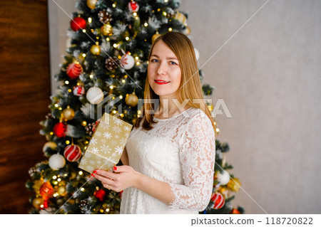 A young woman in a white dress is holding a gift in her hands against Christmas tree 118720822