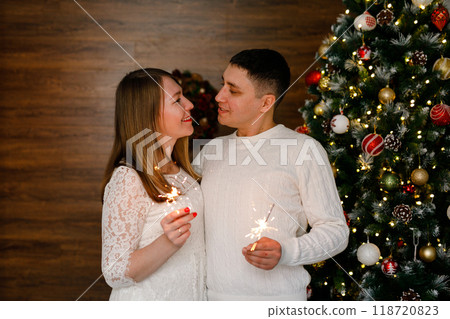 A loving couple is embracing and holding sparklers against the backdrop of a Christmas tree 118720823
