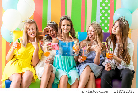 Pretty smiling teenage girls in dresses and crowns sit hugging together holding beverages and taking selfie at birthday party on the striped colorful background 118722333