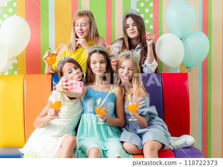 Pretty smiling teenage girls in dresses and crowns sit hugging together holding beverages and taking selfie at birthday party Pretty smiling teenage girls in dresses and crowns sit hugging together holding beverages and taking selfie at birthday party 118722373