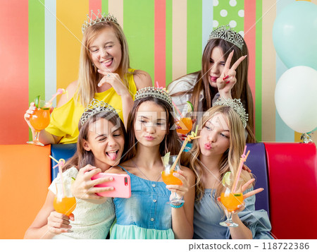 Pretty smiling teenage girls in dresses and crowns sit hugging together holding beverages and taking selfie at birthday party on the striped colorful background 118722386
