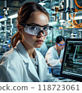 A young female scientist in a white coat and goggles analyzes data on a computer in a modern lab. The focused atmosphere highlights innovation, teamwork, and women in STEM. 118723061