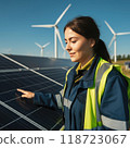 A young woman stands looking at solar panels. Behind her, wind turbines rise against a clear sky, symbolizing women leadership in the green energy transition. 118723067