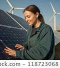 A young woman stands looking at solar panels. Behind her, wind turbines rise against a clear sky, symbolizing women leadership in the green energy transition. 118723068
